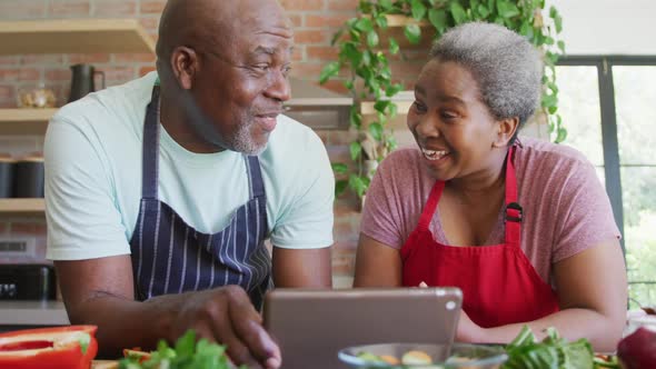 Happy african american senior couple cooking together, using tablet in kitchen