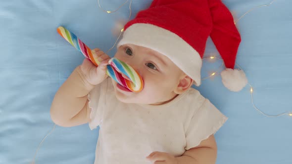 Smiling Funny Newborn Baby in Santa Claus Hat Red Bodysuit Play on White Soft Bed Lick Sweet