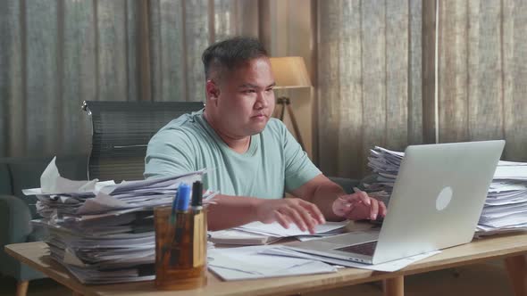 Fat Asian Man Using A Laptop Then Taking Note While Working With Documents At The Office