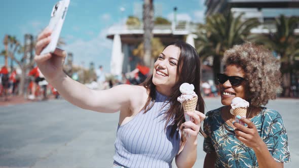Two smiling multiethnic women making selfie with ice cream on phones camera