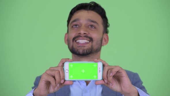 Face of Happy Young Bearded Persian Businessman Thinking While Showing Phone