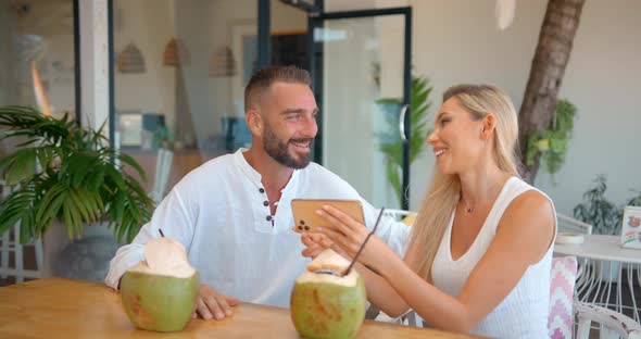 Woman and Man in White Outside at Table with Coconut Cocktails Using Phone Connecting with Friends