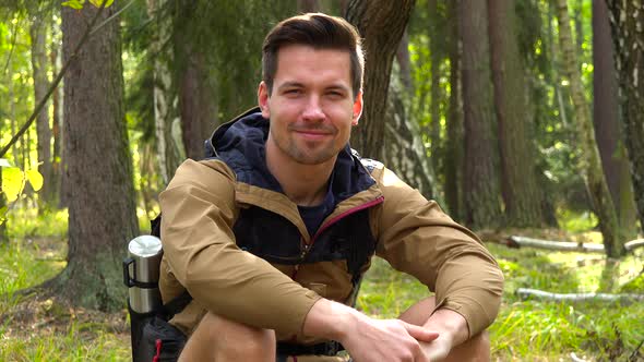 A Young Handsome Hiker Sits on the Ground in a Forest and Smiles at the Camera