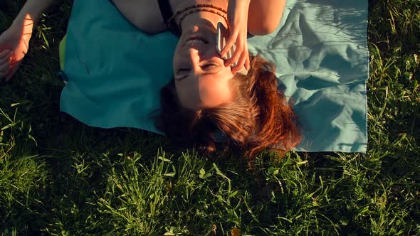 Beautiful Red Hair Young Woman Lying on Grass and Calling on Mobile Phone.