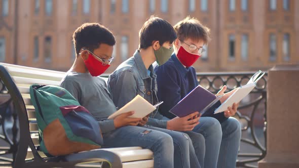 Diverse Schoolmates in Safety Mask Sitting with Books on Bench Outdoors and Studying on Sunny Day.
