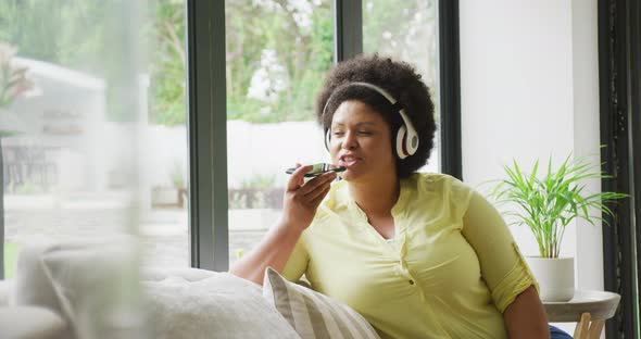 Video of happy plus size african american woman with headphones sitting on sofa with smartphone