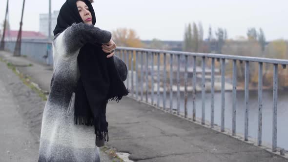 Adult Woman Walks Across the Bridge in Cold Windy Grey Weather and Stopped To Wear Scarf on the Head