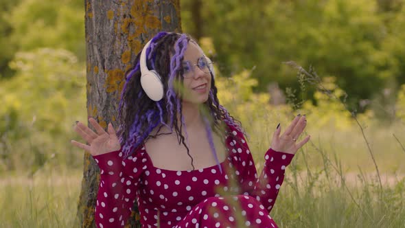 Young Relaxed Woman in Glasses Headphones Sitting Near Tree Listening to Music Dancing in Summertime