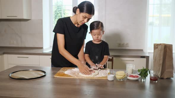 Woman and Girl Stretch Dough on Wooden Board in Kitchen