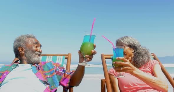 Front view of active senior African American couple toasting drinks on deckchair on the beach 4k