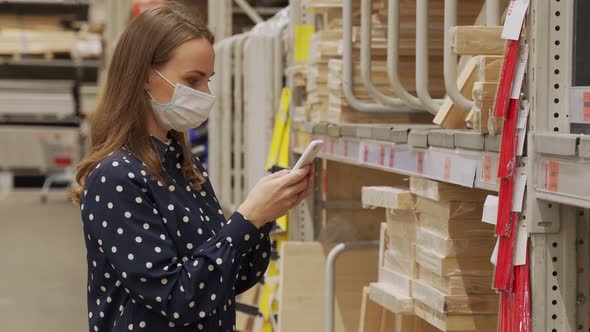 Woman Wearing a Protective Mask in Store Chooses Timber for Carpentry Work Uses Smartphone