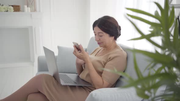 Portrait of Middle-aged Caucasian Woman Sitting with Laptop and Smartphone and Surfing Internet
