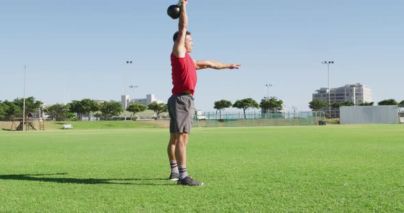 Fit caucasian man exercising outdoors, squatting and lifting kettlebell weight with one arm