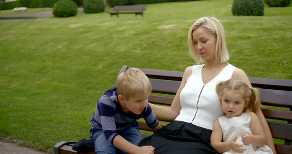 Portrait of the Family: Blonde Mother Sitting on a Bench in the Park and Hugs and Stroking Children