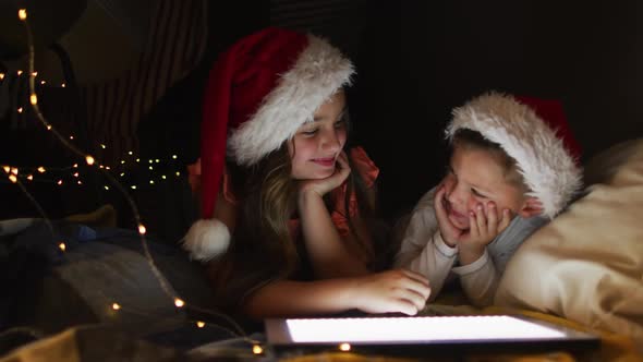 Happy caucasian siblings wearing santa hats lying in makeshift tent, using tablet