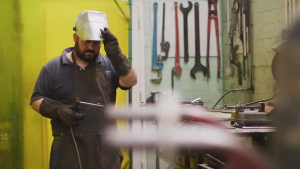 Caucasian male factory worker at a factory making taking off a welding mask, holding welding gun