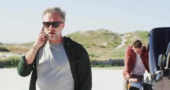 Stressed caucasian gay male couple standing by broken down car, talking on smartphone at the beach