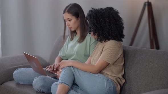 Side View of African American Young Woman Sitting on Couch Helping Asian Roommate with Remote