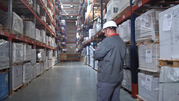 Warehouse Worker Goes Back to the Camera Between the Racks with the Goods