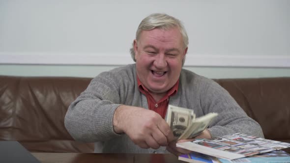 Happy Old Man Throwing Away the Dollars Sitting at the Wooden Table Close Up. Positive Rich Man