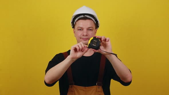 Young Handsome Builder in a Hardhat Holds a Tape Measure Ruler in His Hands on a Yellow Background