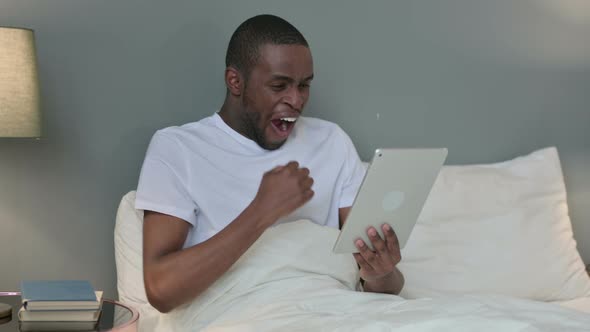 Excited African Man Celebrating on Tablet in Bed 