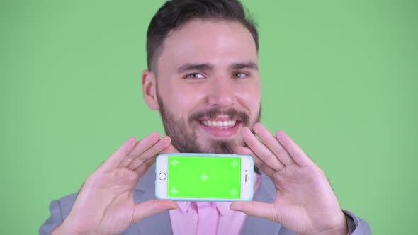 Face of Happy Young Bearded Businessman Thinking While Showing Phone