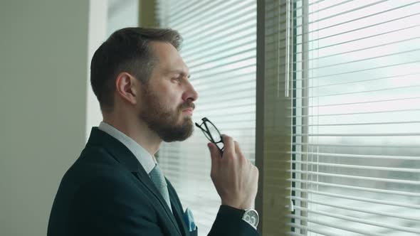 Focused Businessman in a Suit Stands at the Panoramic Window in the Office and Reflects the Thought