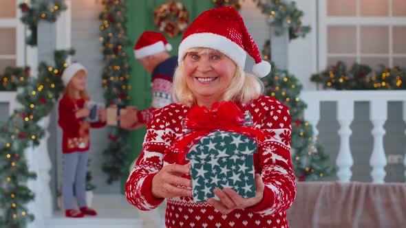 Portrait of Grandmother Woman Presenting Gift Box Smiling Near Decorated Christmas House with Family