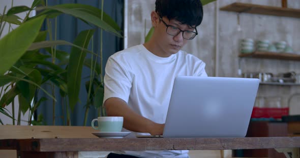 Man Asian in Glasses a Teenager, Working on a Laptop Looks at the Screen While Sitting in a Cafe