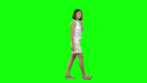 Young Woman with Brown Haircut Is Calmly Walking and Talking on Mobile Phone on Greenscreen. Profile