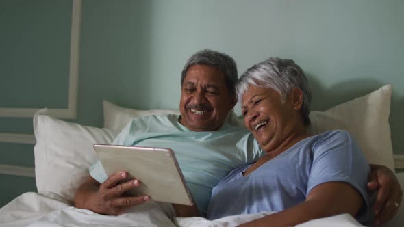 Happy senior mixed race couple sitting in bed using tablet