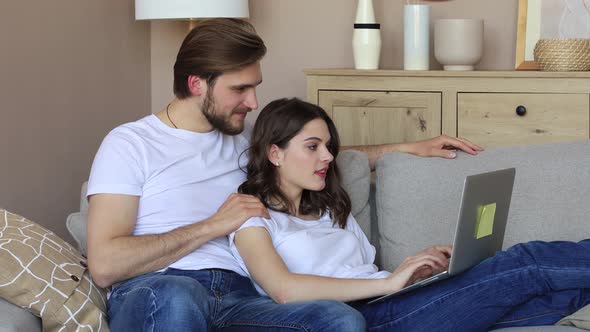 Happy couple sitting, relaxing on sofa in living room, using laptop for online shopping together