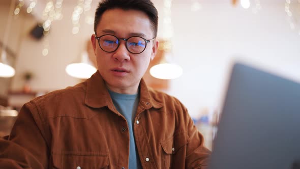 Pensive Asian young man wearing eyeglasses working on laptop