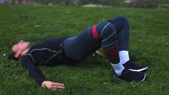 a Girl in Blue Leggings Lying on Green Grass and Doing Exercises with Rubber for the Buttocks