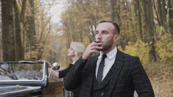 Portrait of Gentleman Stands in Front of His Lady and Smokes Cigar in Forest