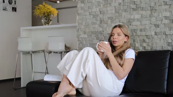 Young blond woman drinks tea on the couch at home and thinking about something