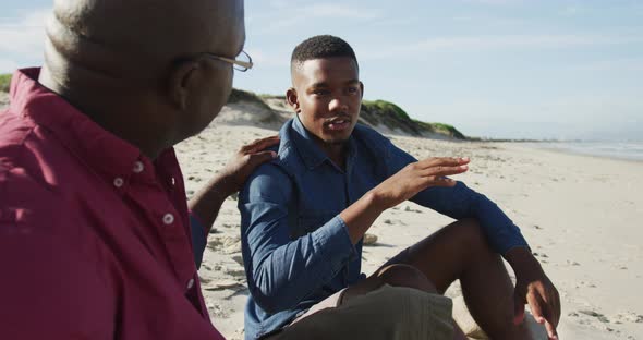 African american father sitting on beach with teenage son, putting hand on his shoulder and talking