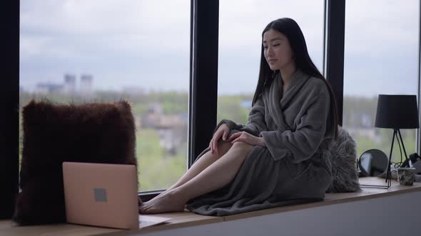 Wide Shot of Gorgeous Asian Young Woman Sitting on Windowsill Smiling at Laptop Web Camera