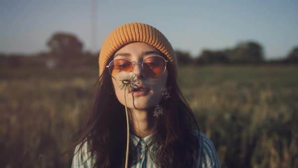 A Beautiful Young Woman in Stylish Sunglasses Stand in a Field and Blow a Dandelion