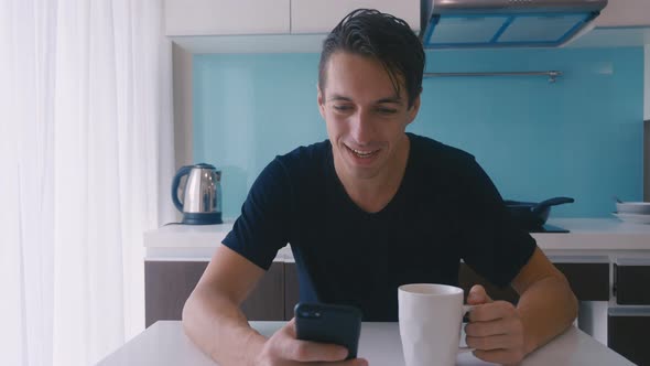 Young Smiling Man Using Smartphone and Drinking Coffee at Home at Kitchen