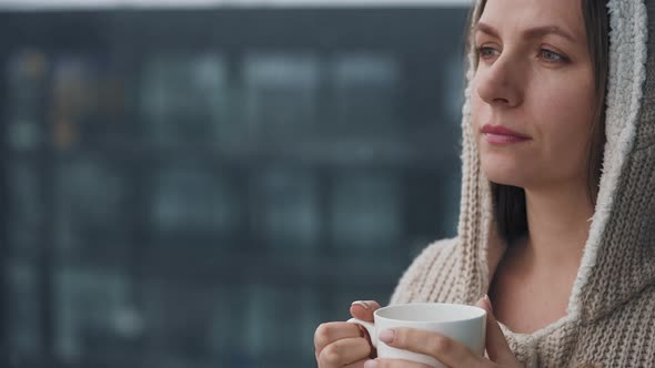 Caucasian Woman Stays on Balcony During Snowfall with Cup of Hot Coffee or Tea