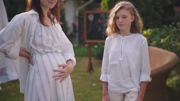 Portrait of Unsatisfied Caucasian Teenage Girl Standing with Unrecognizable Pregnant Mother on