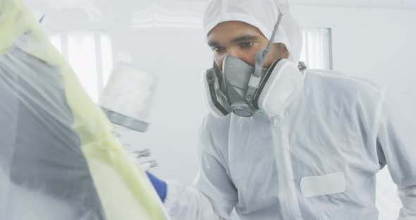 Mixed race male car mechanic with protective clothes and using a spraying gun on a piece of a car