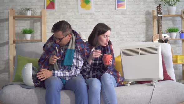 Frozen Young Woman and Man Wrapped in a Blanket in the Living Room Are Heated Next To Electric