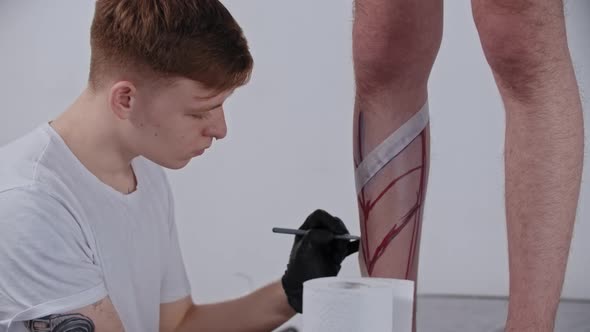 Young Man Tattoo Artist Drawing a Sketch on the Leg on His Client with a Marker