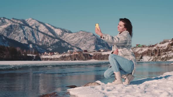 A Woman Tourist Sits on the River Bank in a Mountainous Area and Takes a Selfie Using a Smartphone