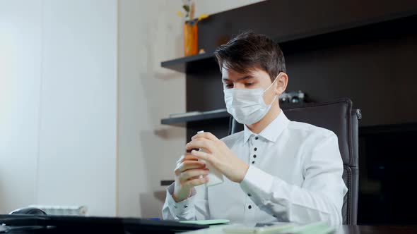 A Young Man in a Medical Mask Sitting at the Table Disinfects His Hands