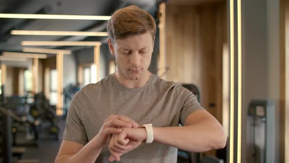 Smiling Bodybuilder Using Smart Watch at Gym. Sportsman Standing in Fitness Club alt