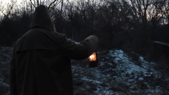 a Man in a Dark Cloak Walks with a Lantern in a Dusky Forest in the Dark